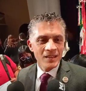 Persona en un evento público, con fondo de varias personas y banderas, usando traje formal y haciendo un gesto con la cara.