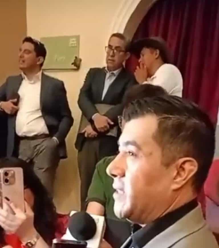 Persona hablando en un evento formal con varias personas en el fondo, algunos en conversación y otros escuchando atentamente.