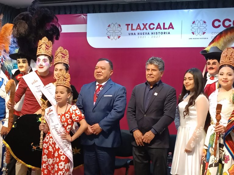 Grupo de personas en evento cultural en Tlaxcala, con personajes en trajes tradicionales y disfraces, en un escenario con cartel de "Tlaxcala, una nueva historia".