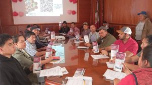 Capacitación y discusión en una sala de reuniones con varios participantes, algunos con folletos y laptops, en un entorno formal de trabajo.