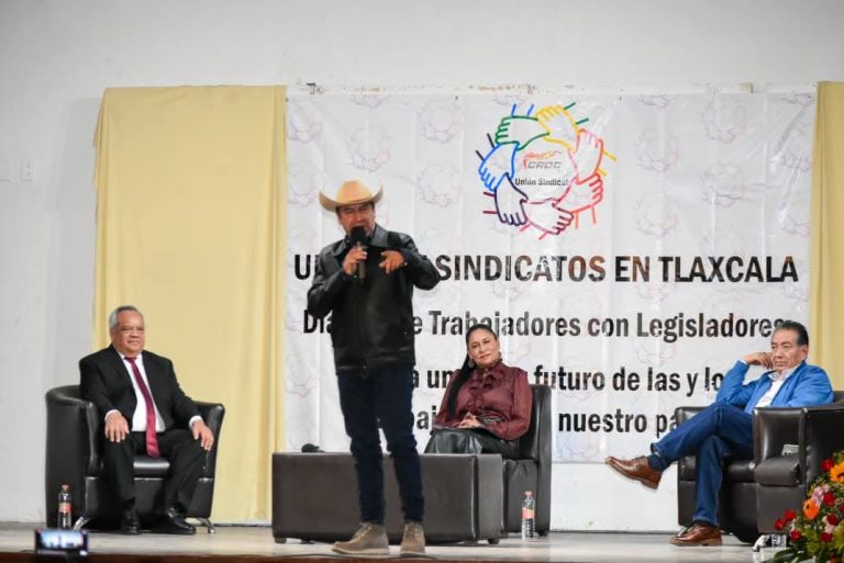 Hombre con sombrero y chaqueta negra hablando en un escenario durante un evento con tres personas sentadas en sillones.