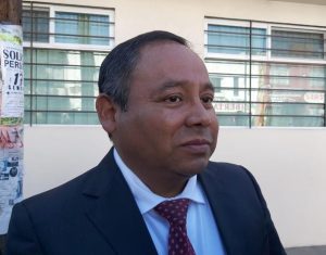 Hombre con traje y corbata en un entorno urbano, posiblemente en una calle o cerca de un edificio, con ventanas y carteles en el fondo.