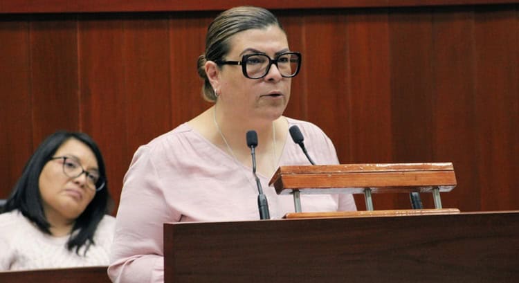 Mujer en un podio hablando en un entorno formal, posiblemente en una conferencia o audiencia oficial.