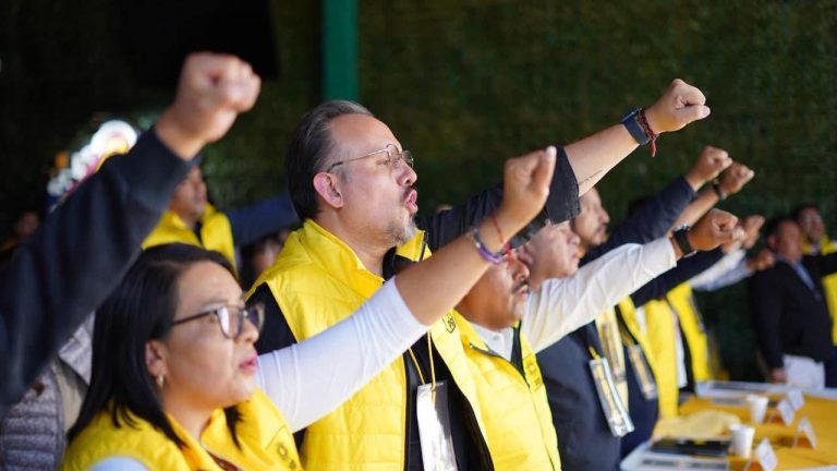 Líderes y participantes en una ceremonia con gestos de protesta, usando chaquetas amarillas, en un entorno al aire libre con árboles de fondo.
