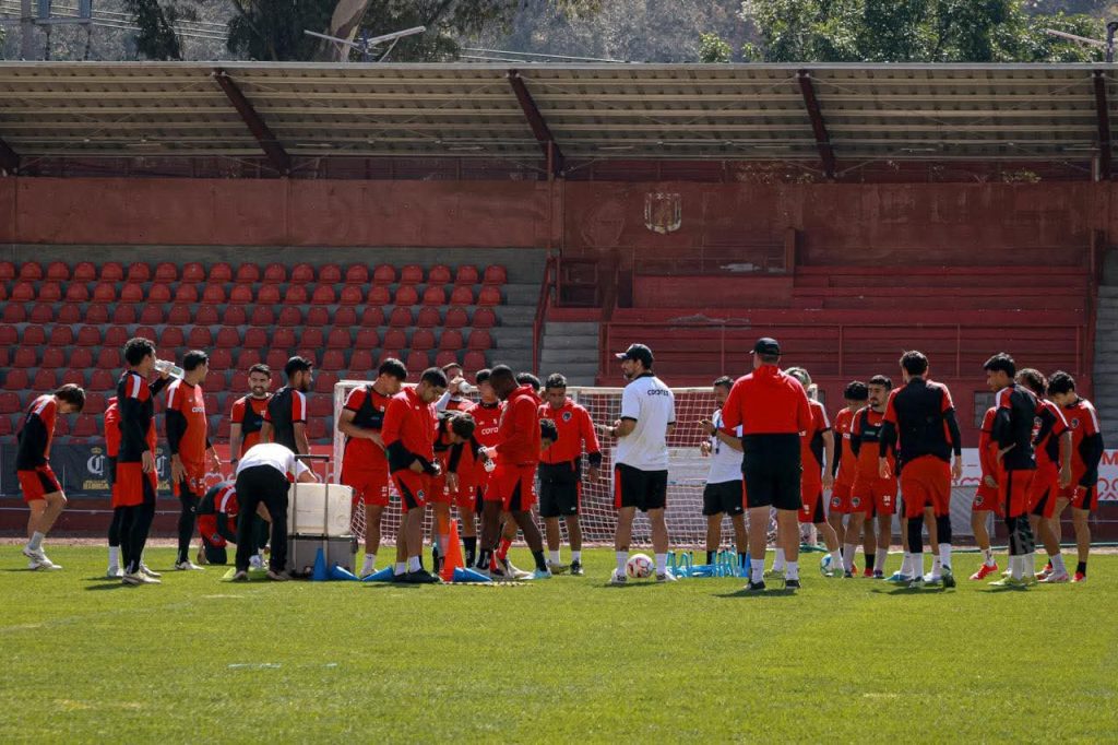 Equipo de fútbol en entrenamiento en un campo de césped, con jugadores y entrenadores en una sesión de práctica en un estadio con gradas rojas.
