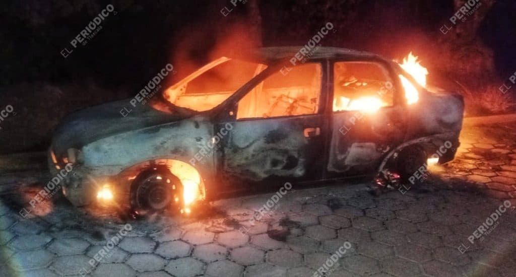 Incendio de un coche en la calle durante la noche, con llamas y humo saliendo del vehículo, en un entorno oscuro y desolado.