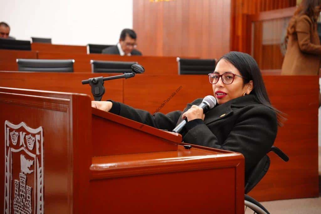 Mujer en silla de ruedas hablando en un micrófono en un entorno formal, con fondo de madera y otras personas en un espacio de reunión o conferencia.