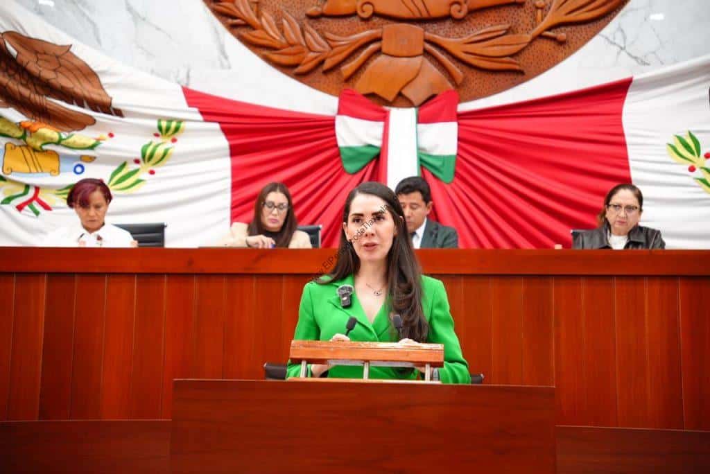 Mujer en un podio en una sesión del Congreso de Tlaxcala, con fondo decorativo con bandera y escudo del estado.