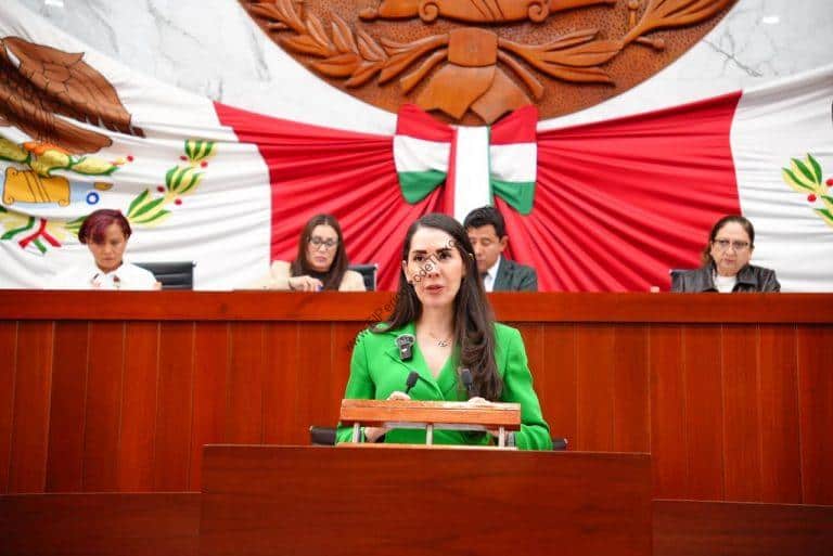Mujer en un podio en una sesión del Congreso de Tlaxcala, con fondo decorativo con bandera y escudo del estado.