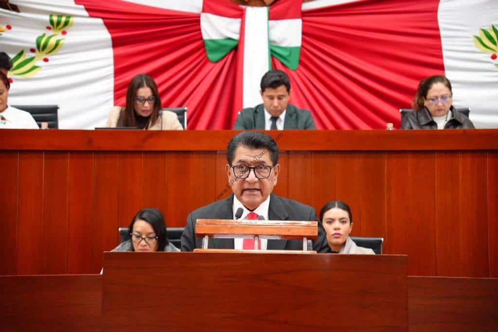 Diputado en sesión del Congreso de Tlaxcala con fondo de bandera mexicana y decoraciones patrióticas.