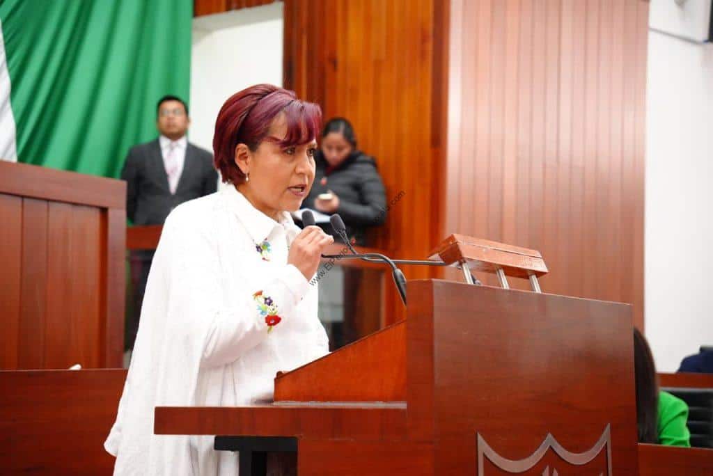 Mujer en un podio hablando en un evento formal en un salón con paredes de madera y fondo con cortina verde.