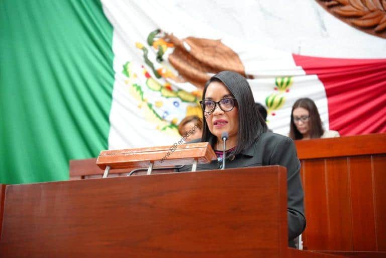 Mujer hablando en un podio con bandera de México de fondo, en un evento oficial.