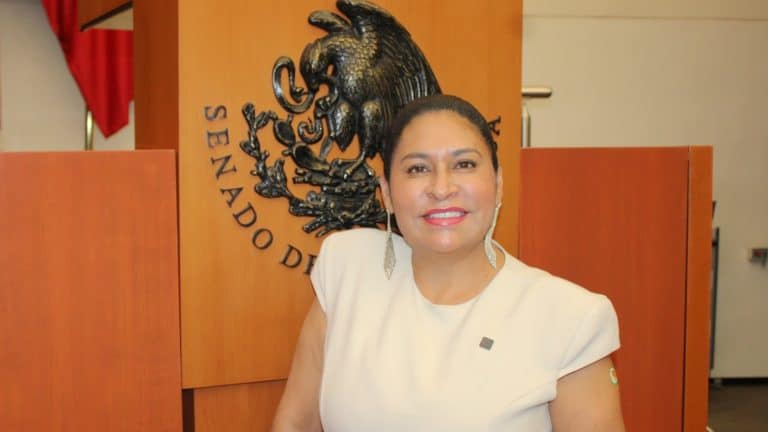 A woman smiling in front of a wooden panel with the emblem of the Senate of Mexico, wearing a white dress and earrings, in an official setting.