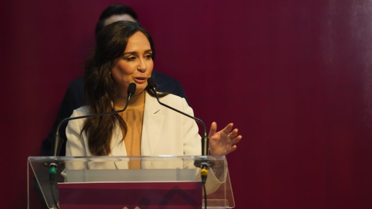 Mujer hablando en un podio durante una conferencia o evento público, con fondo de color burdeos.