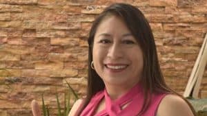 Mujer sonriendo con blusa rosa en un entorno con pared de ladrillos y plantas verdes.