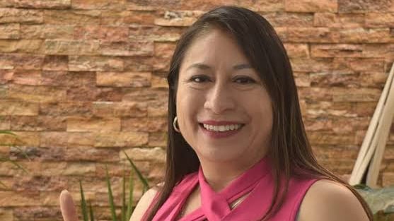 Mujer sonriendo con blusa rosa en un entorno con pared de ladrillos y plantas verdes.