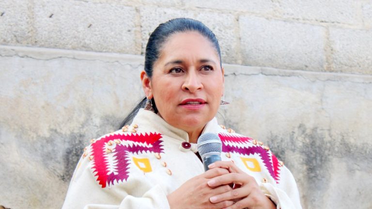 Mujer hablando en un micrófono frente a una pared de piedra, participando en un evento público en Tlaxcala, México.