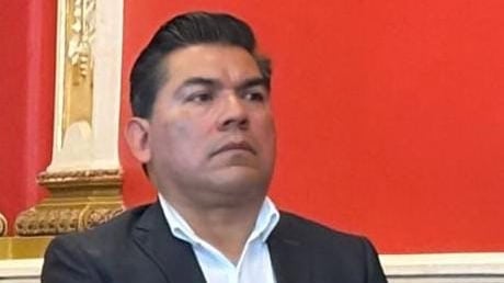 Hombre con cabello oscuro peinado hacia atrás, vestido con saco negro y camisa blanca, en un entorno con fondo rojo y detalles dorados.