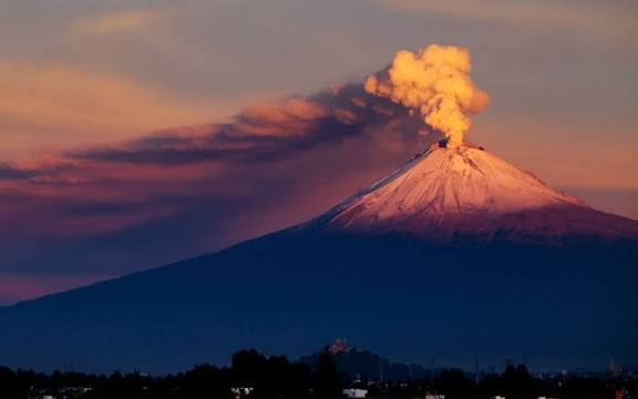 Volcán en erupción con columna de humo y lava al atardecer, en un paisaje natural impresionante.