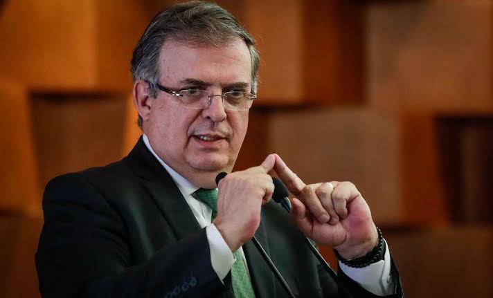 Hombre de negocios con gafas y traje oscuro, hablando en un evento formal, gesticulando con las manos mientras explica un punto importante.