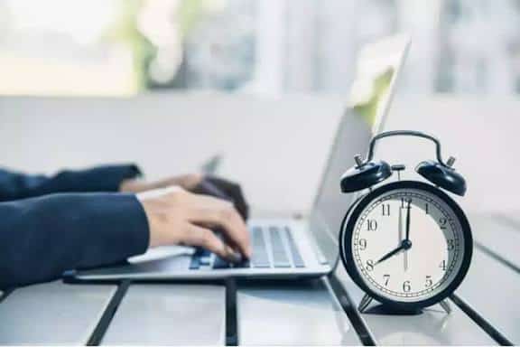 Reloj despertador clásico en una mesa de oficina junto a una persona usando una laptop, simbolizando la gestión del tiempo y la productividad laboral.