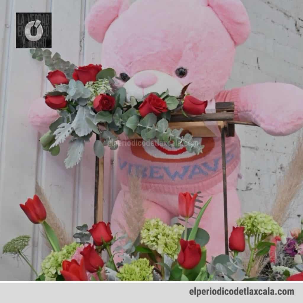 Osito de peluche rosa con ramo de rosas rojas y flores verdes, decorando un espacio con fondo de pared blanca y detalles en tonos pastel.