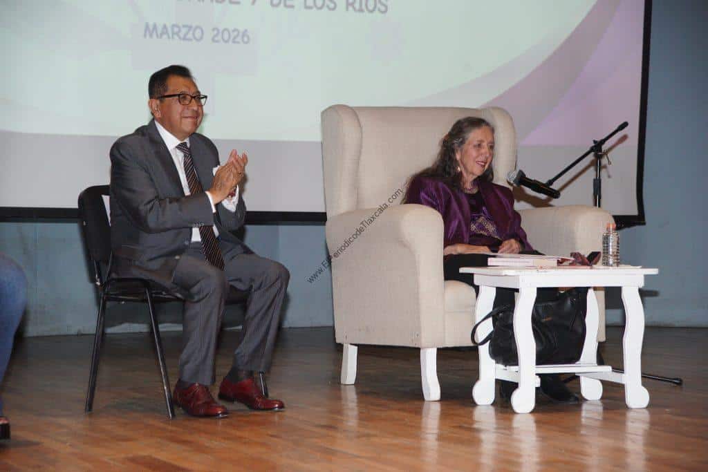 Una conferencia con un hombre y una mujer en un escenario, la mujer está sentada en un sillón grande y la otra persona en una silla, ambos parecen participar en un evento formal o académico.