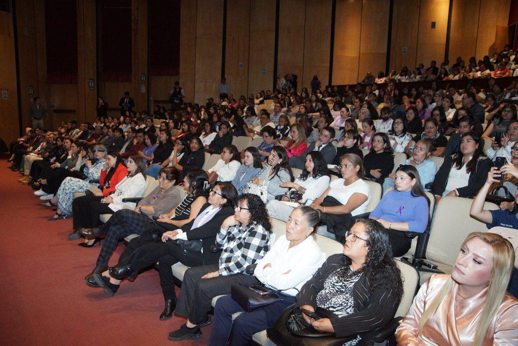 Asistentes en un auditorio durante un evento o conferencia, sentados en filas y prestando atención a la presentación. La sala está llena de personas de diferentes edades y estilos, participando en una.