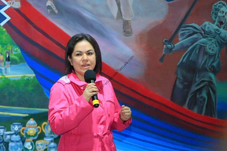 Una mujer con chaqueta rosa hablando en un micrófono frente a un mural colorido que representa un barco y figuras históricas.