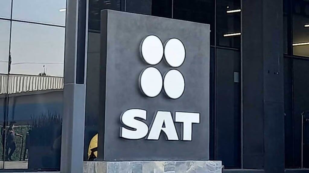 SAT logo en un edificio moderno con fachada de vidrio y estructura metálica, en color blanco sobre fondo gris oscuro.