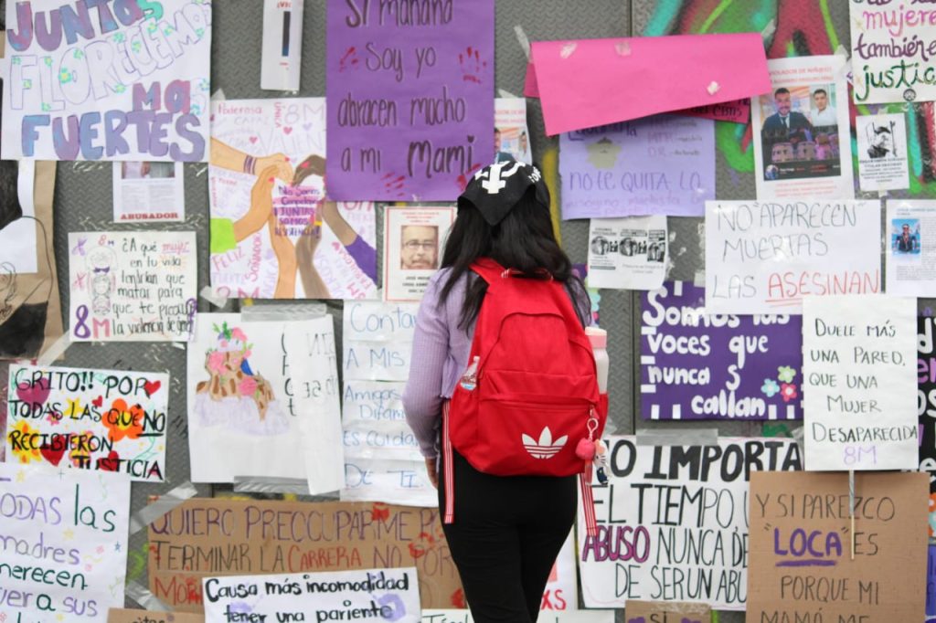 Mujer con mochila roja frente a un mural de carteles y mensajes en protesta por violencia de género y justicia en Tlaxcala, México.