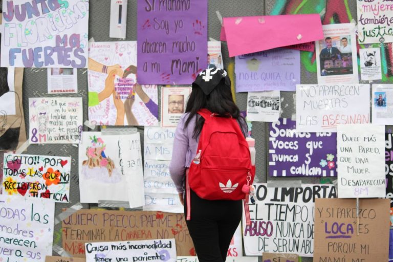 Mujer con mochila roja frente a un mural de carteles y mensajes en protesta por violencia de género y justicia en Tlaxcala, México.