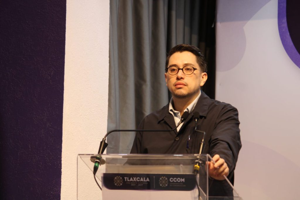Un hombre con gafas y chaqueta negra hablando en un podio durante una conferencia o evento formal en Tlaxcala, México.