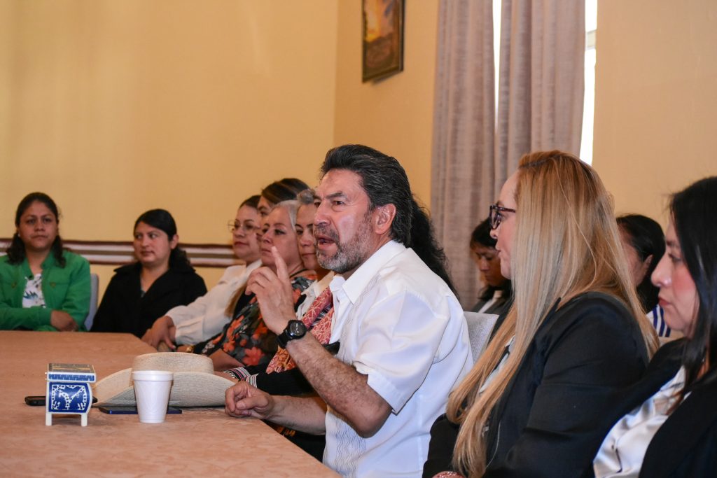 Un grupo de personas participa en una reunión formal en una sala con cortinas y cuadros en las paredes, destacando un hombre en el centro que habla y gesticula durante la discusión.
