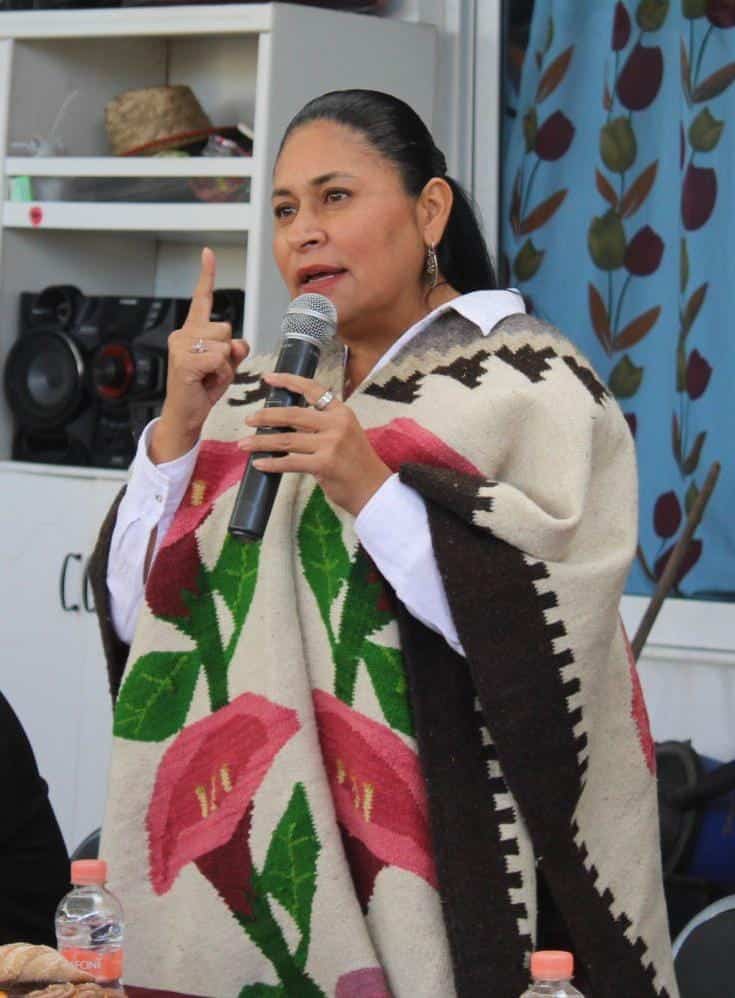 Mujer hablando en un evento, usando un poncho con diseño floral y sosteniendo un micrófono, en un entorno interior con estantería y decoraciones en el fondo.