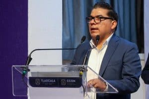 Un hombre con gafas y traje azul hablando en un podio durante una conferencia o evento oficial en Tlaxcala, México.