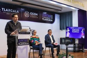 Panel de discusión en evento de comunicación en Tlaxcala con tres participantes y un presentador, en un escenario con pantallas y decoraciones oficiales.