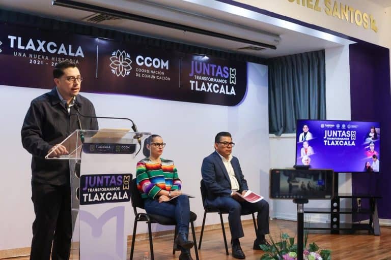 Panel de discusión en evento de comunicación en Tlaxcala con tres participantes y un presentador, en un escenario con pantallas y decoraciones oficiales.