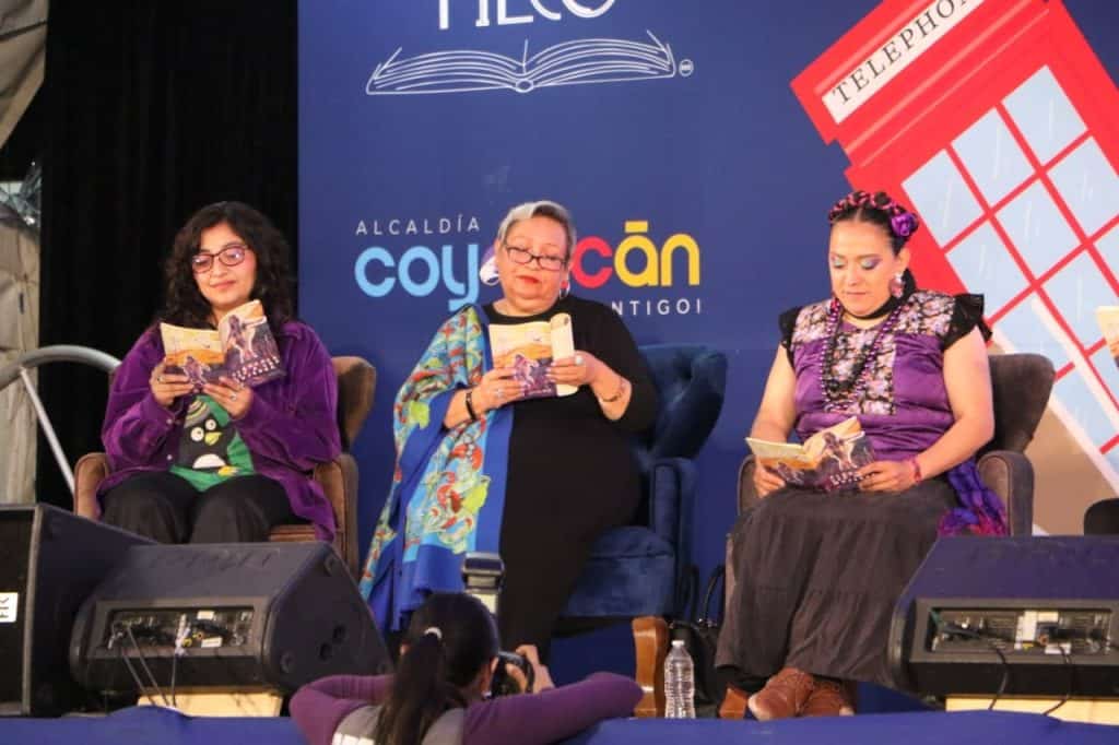 Tres mujeres en un panel de discusión leyendo libros en un evento cultural, con un fondo que incluye un logo de la alcaldía de Coyoacán y una imagen de una cabina telefónica roja.