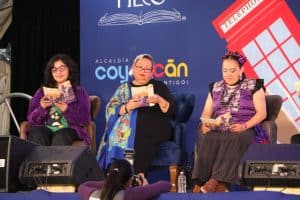 Tres mujeres en un panel de discusión leyendo libros en un evento cultural, con un fondo que incluye un logo de la alcaldía de Coyoacán y una imagen de una cabina telefónica roja.