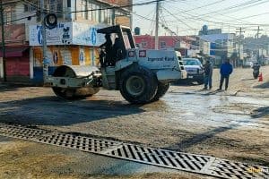 Máquina compactadora de pavimento trabajando en una calle de Tlaxcala, en un día soleado, con edificios y personas observando en el fondo.