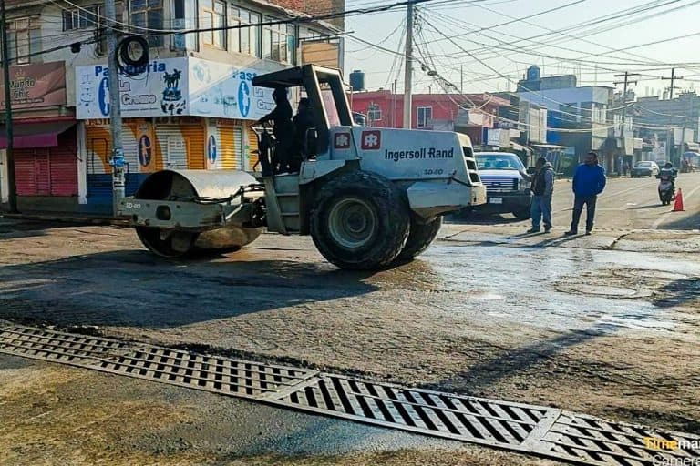 Máquina compactadora de pavimento trabajando en una calle de Tlaxcala, en un día soleado, con edificios y personas observando en el fondo.
