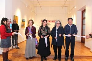 Personas en una inauguración de exposición de arte en una galería, cortando la cinta de apertura, con obras y espectadores en el fondo.