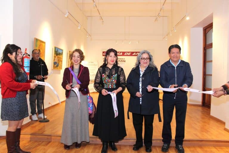Personas en una inauguración de exposición de arte en una galería, cortando la cinta de apertura, con obras y espectadores en el fondo.
