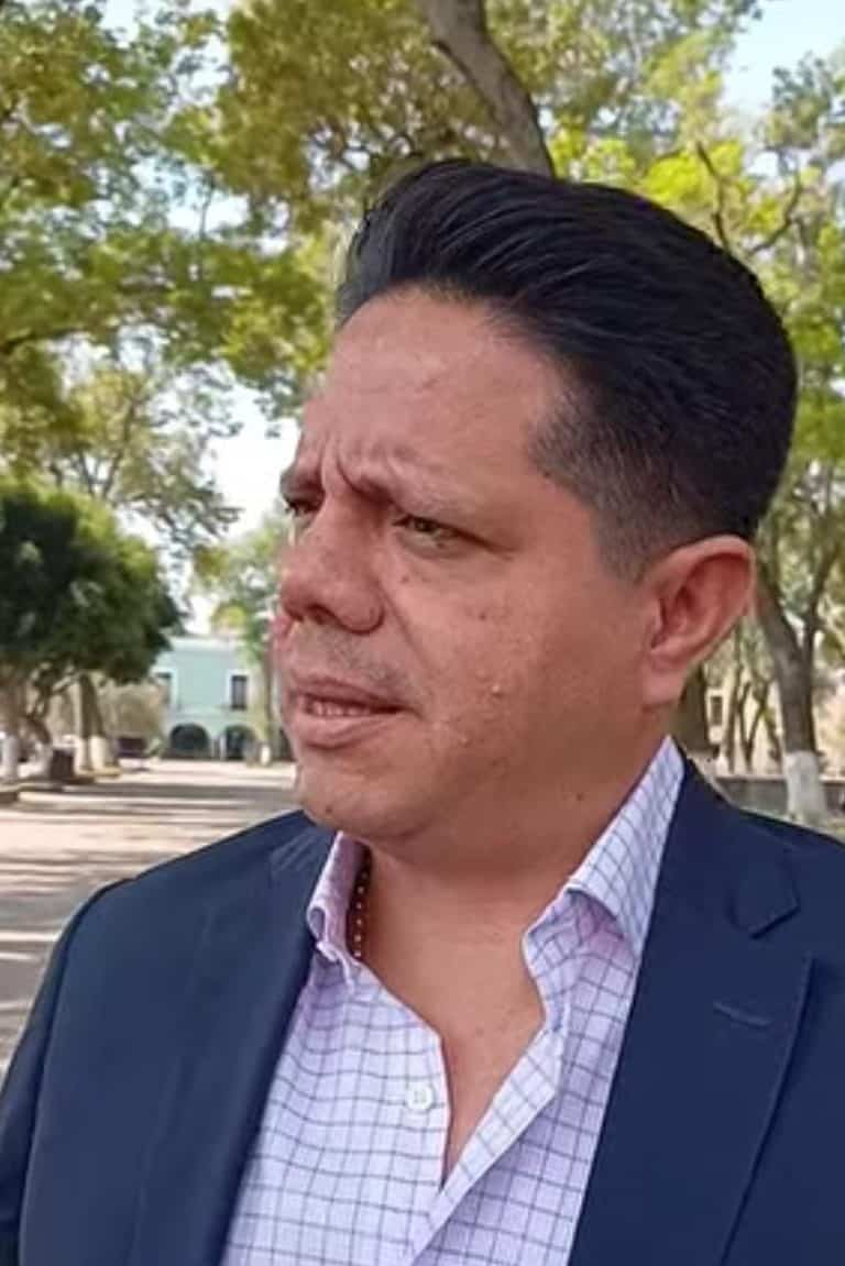 Retrato de un hombre con peinado peinado hacia atrás, usando chaqueta azul y camisa a cuadros, en un entorno al aire libre con árboles y edificios en el fondo.