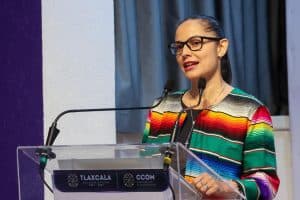 Una mujer con gafas y cabello recogido, vestida con un traje tradicional colorido, hablando en un podio durante una conferencia o evento oficial en Tlaxcala, México.