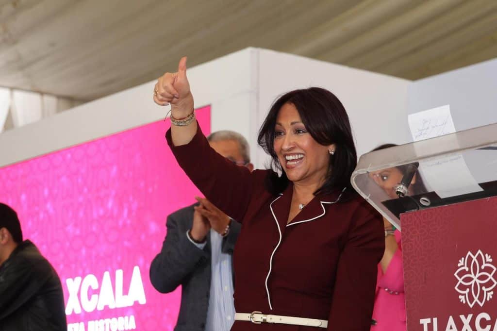 Voz femenina en evento público, sonriendo y haciendo señal de aprobación con el pulgar hacia arriba, en un entorno con fondo rosa y personas en el fondo.