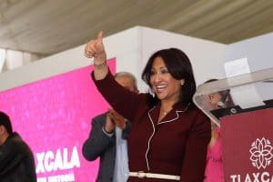 Voz femenina en evento público, sonriendo y haciendo señal de aprobación con el pulgar hacia arriba, en un entorno con fondo rosa y personas en el fondo.
