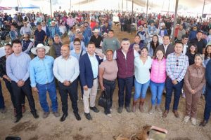 Amplia vista de un evento público con un grupo de personas en primer plano, incluyendo a autoridades y asistentes, en un espacio cubierto con muchas personas participando en la actividad.