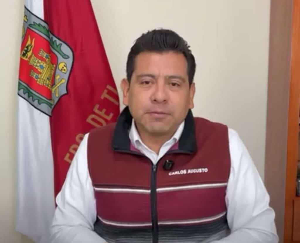 Persona en una oficina con bandera de Tlaxcala al fondo, usando un chaleco con su nombre, en un entorno formal y profesional.
